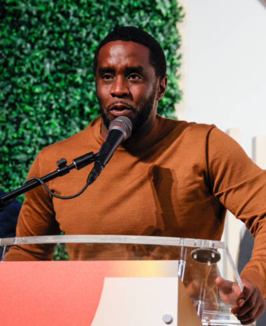 Diddy stands behind a clear podium, speaking into a microphone while wearing a brown long-sleeved shirt, framed by a lush green leafy backdrop.