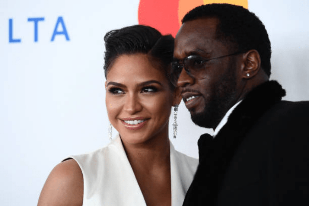 Diddy wears a black fur coat with a left-ear earring, standing beside Cassie in a sleeveless white outfit at an event with a "DELTA" sign and a red-orange circular logo in the background.