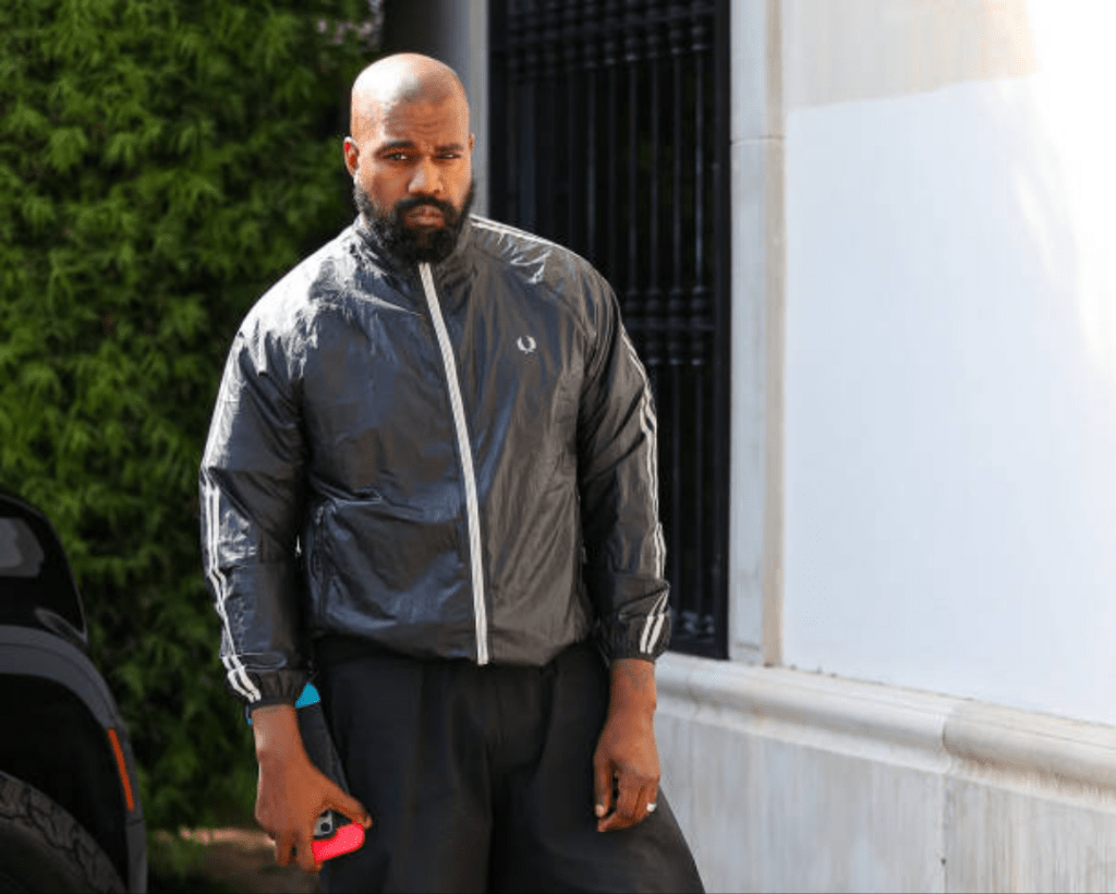 Kanye West stands confidently in stylish attire, his expression intense and focused, set against a minimalist backdrop that emphasizes his iconic presence.