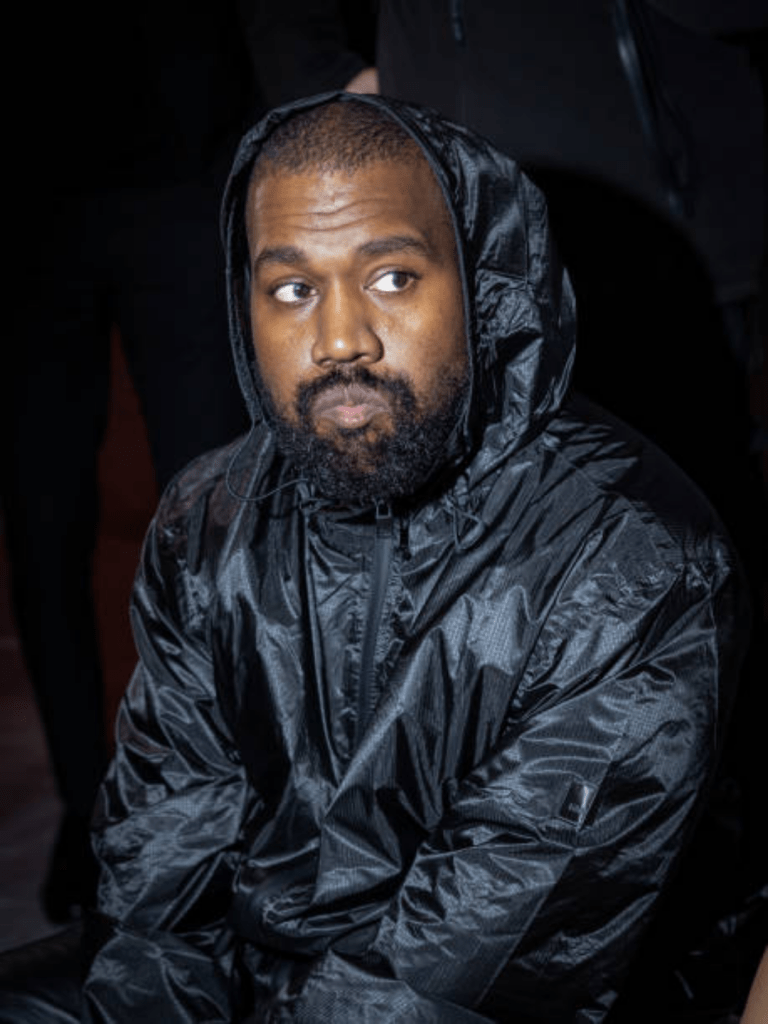 Kanye West wears a black, shiny, hooded jacket with a textured, waterproof look, standing against a dark backdrop with partially visible figures behind him.