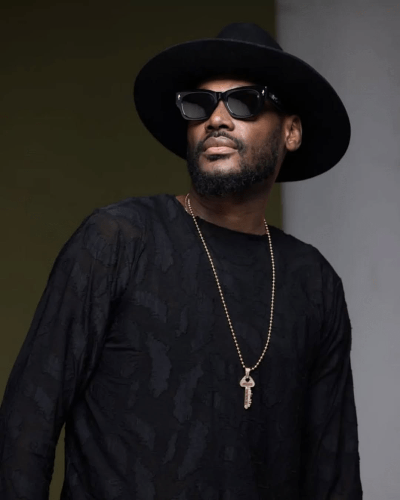 Singer 2Baba wears a black hat and patterned shirt, accessorized with a gold chain and key pendant, set against a dark green and light grey background.