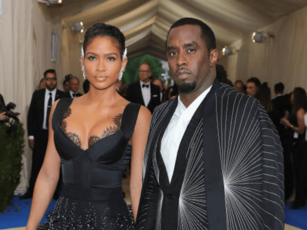 Diddy wears a black suit with a white shirt and a dramatic pinstriped cape, while Cassie stuns in a black dress with lace detailing and a sparkling skirt at a high-profile formal event.