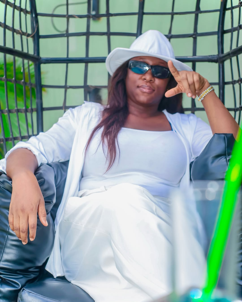 Cynthia Morgan sits in a black cushioned chair with a grid frame, dressed in a white outfit and matching hat, accessorized with a gold bracelet. A green wall and surrounding foliage create a relaxed, stylish backdrop.