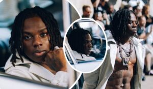 Singer Rema showcases bold style in a white jacket layered with necklaces, including a large pendant. The collage highlights moments from a public setting: him seated in a car, a close-up of his ring-adorned hand, and a striking shirtless stance revealing tattoos and jewelry. The backdrop teems with onlookers, amplifying the sense of star power.