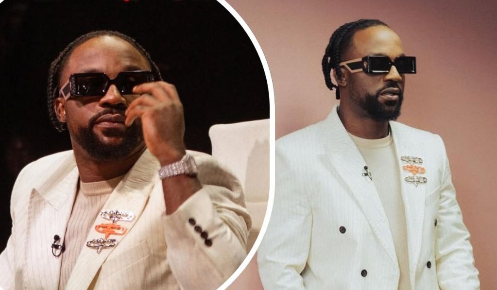 Singer Iyanya dons a sleek cream suit with a double-breasted design and dark buttons, accented by three decorative brooches. His look is completed with a matching shirt, a gleaming wristwatch, and neatly braided hair, set against contrasting backdrops of dark and peach tones.