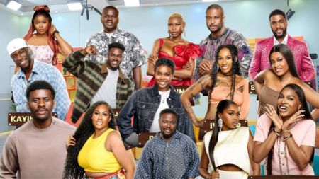 A group photo of BBNaija Season 10 housemates posing inside the Big Brother Naija house. The lineup features both male and female contestants in vibrant outfits, smiling and standing together in the well-lit lounge area.