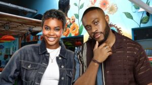 BBNaija Season 10 housemates Dede and Lola posing inside the Big Brother Naija house. Dede is smiling in a denim jacket, while Lola stands beside her in a dark striped shirt with his hand on his chin. A vibrant floral mural serves as the background.