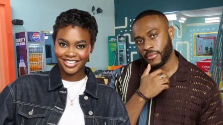 Two individuals, Dede and Kola, stars of BBNaija 10, posing together in a colorful room with vending machines featuring Pepsi and Colgate branding in the background. Dede is wearing a denim jacket over a white top, while Kola is dressed in a brown textured shirt with a thoughtful pose.
