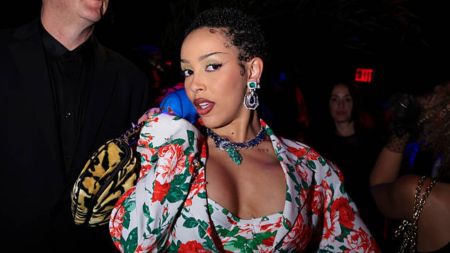 Doja Cat poses in a bold floral outfit with red and green rose prints, accessorised with statement emerald jewellery, at a nightlife event.
