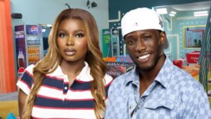 Big Brother Naija season 10 housemates Isabella and Kayikunmi posing together inside the colourful Big Brother house, with branded drinks and the Colgate game table visible in the background.