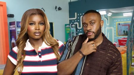 Big Brother Naija Season 10 housemates Isabella and Kola pose inside the BBNaija house. Isabella wears a striped red and white polo shirt with long wavy hair, while Kola poses in a brown striped shirt with his hand on his chin.