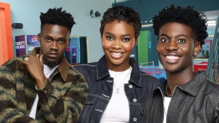 BBNaija Season 10 housemates Jason Jae, Dede, and Koyin posing inside the Big Brother Naija house, with Jason Jae in a patterned green and brown jacket, Dede in a dark denim jacket over a white top, and Koyin in a dark denim jacket, all smiling towards the camera except Jason Jae who has a serious expression.