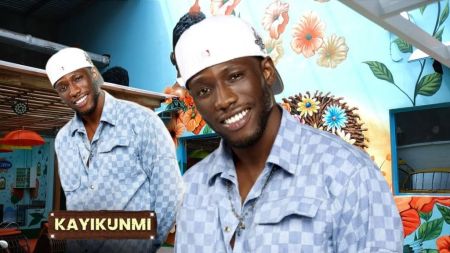 BBNaija Season 10 housemate Kayikunmi smiling in a blue checkered shirt and white cap, with a floral mural background inside the Big Brother house. A second image of him with a name tag is placed beside him.