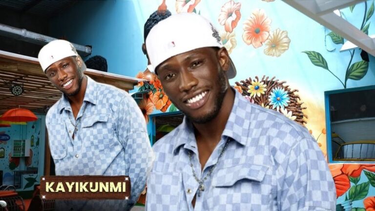 BBNaija Season 10 housemate Kayikunmi smiling in a blue checkered shirt and white cap, with a floral mural background inside the Big Brother house. A second image of him with a name tag is placed beside him.
