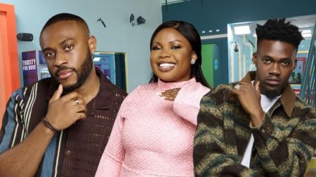 BBNaija Season 10 housemates Kola, Doris, and Jason Jae posing inside the Big Brother Naija house, with Kola in a patterned brown shirt, Doris in a pink patterned long-sleeve dress, and Jason Jae in a green-and-brown checkered jacket over a white shirt.