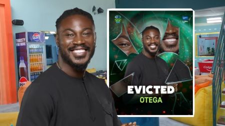 BBNaija Season 10 housemate Otega smiling in the Big Brother Naija house, with his official eviction card showing "Evicted Otega" placed beside him.