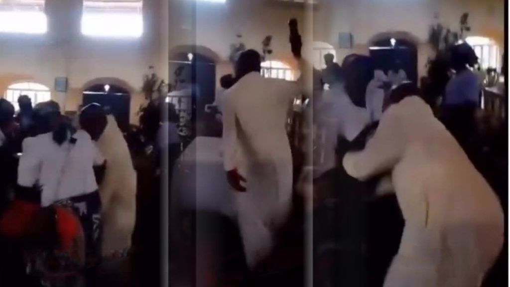 Inside a church, a priest in a white robe is seen confronting and physically halting women during what appears to be a Catholic women’s August meeting, with several attendees present and watching.
