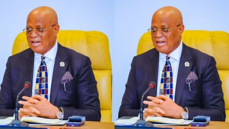 Akwa Ibom State Governor seated at a meeting table, wearing a dark suit, patterned tie, white shirt, and glasses, speaking into a microphone with a notepad and pen in front of him.
