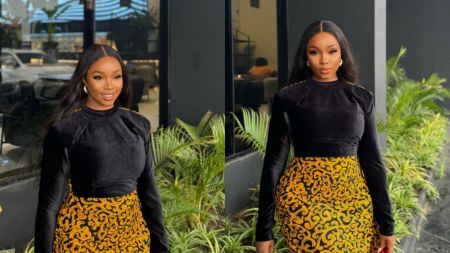 Nigerian reality TV star Bambam poses outdoors in a stylish outfit, wearing a black long-sleeved velvet top paired with a fitted golden patterned skirt. She has long wavy hair, hoop earrings, and a bright smile.