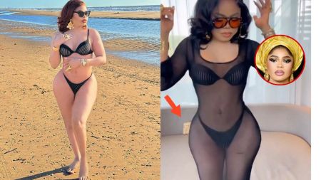 Bobrisky stands indoors in a sheer black mesh dress over a black bikini, hands raised slightly, wearing large sunglasses and gold bracelets in front of a beige sofa; a red arrow points to the sofa while a circular inset at top right shows Bobrisky’s headshot in a gold gele.