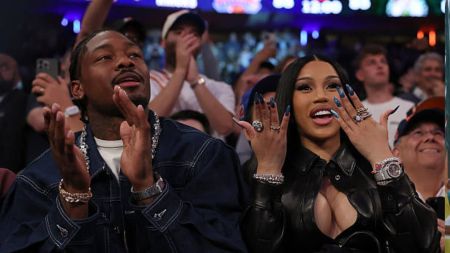American football star Stefon Diggs and rapper Cardi B sit courtside at a basketball game, smiling and clapping. Cardi B shows off her long blue nails and diamond rings while Diggs applauds. Both wear stylish outfits and jewellery, with cheering fans visible in the background.