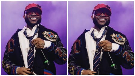 Nigerian singer Davido performs on stage, smiling in a red cap, dark sunglasses, and a stylish black jacket with crest embroidery, paired with a white shirt and striped tie.