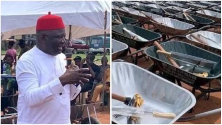 Anambra lawmaker Dominic Okafor speaking at an event distributing branded wheelbarrows and cutlasses to community members, with recipients seated under tents in the background.