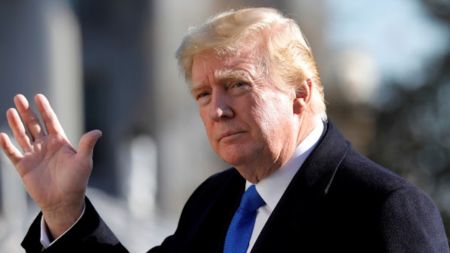 Donald Trump raises his hand while wearing a dark coat and blue tie, looking straight ahead with a serious expression.