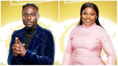 BBNaija Season 10 housemates Faith and Doris pose against a bright yellow backdrop. Faith is dressed in a deep blue velvet jacket with his hands clasped, while Doris wears a pink long-sleeve textured dress, smiling warmly at the camera.