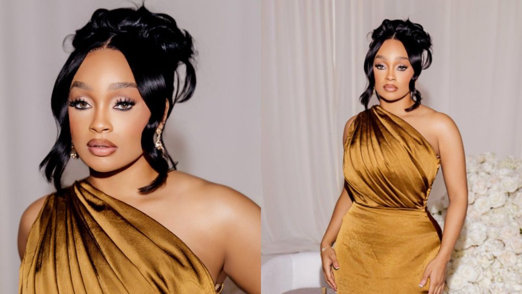 Nigerian actress Folasade poses in an elegant one-shoulder golden gown, styled with statement earrings and soft glam makeup, standing against a neutral background with white floral décor.