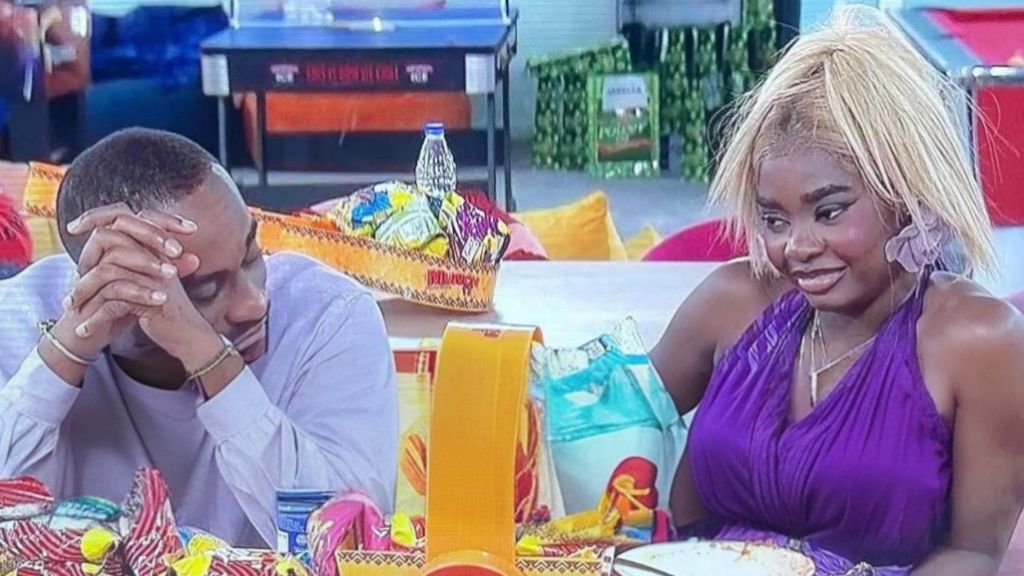 BBNaija Season 10 housemates Kola and Dede sitting together at the dining table, with Kola resting his head on folded hands while Dede, dressed in a purple outfit, looks at him with a smile.