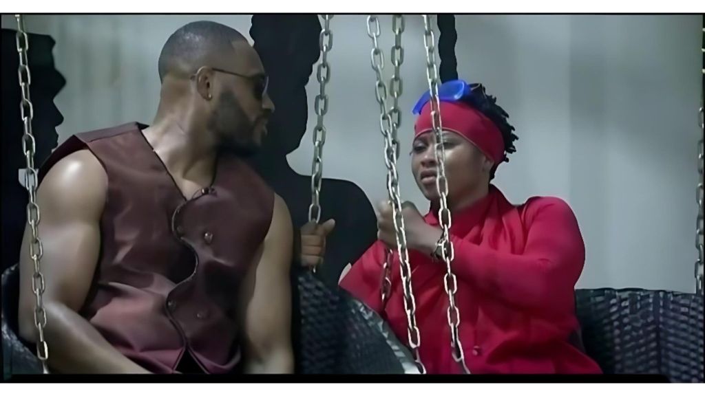 BBNaija Season 10 housemates Kola and Imisi sit on a swing set indoors, engaged in a close conversation. Kola wears a sleeveless brown outfit and sunglasses, while Imisi is dressed in a bright red outfit with a matching headband and blue goggles resting on her head.
