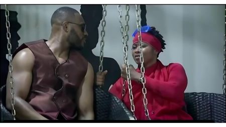 BBNaija Season 10 housemates Kola and Imisi sit on a swing set indoors, engaged in a close conversation. Kola wears a sleeveless brown outfit and sunglasses, while Imisi is dressed in a bright red outfit with a matching headband and blue goggles resting on her head.