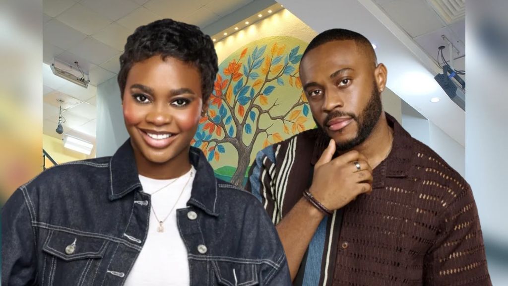 Dede and Kola, housemates from BBNaija Season 10, stand side-by-side indoors. Dede, in a dark denim jacket over a white top with a short pixie cut, smiles toward the camera. Kola, in a brown knit shirt with striped panels, rests his hand on his chin. Behind them is a brightly lit hallway with a colorful tree mural and ceiling lights.