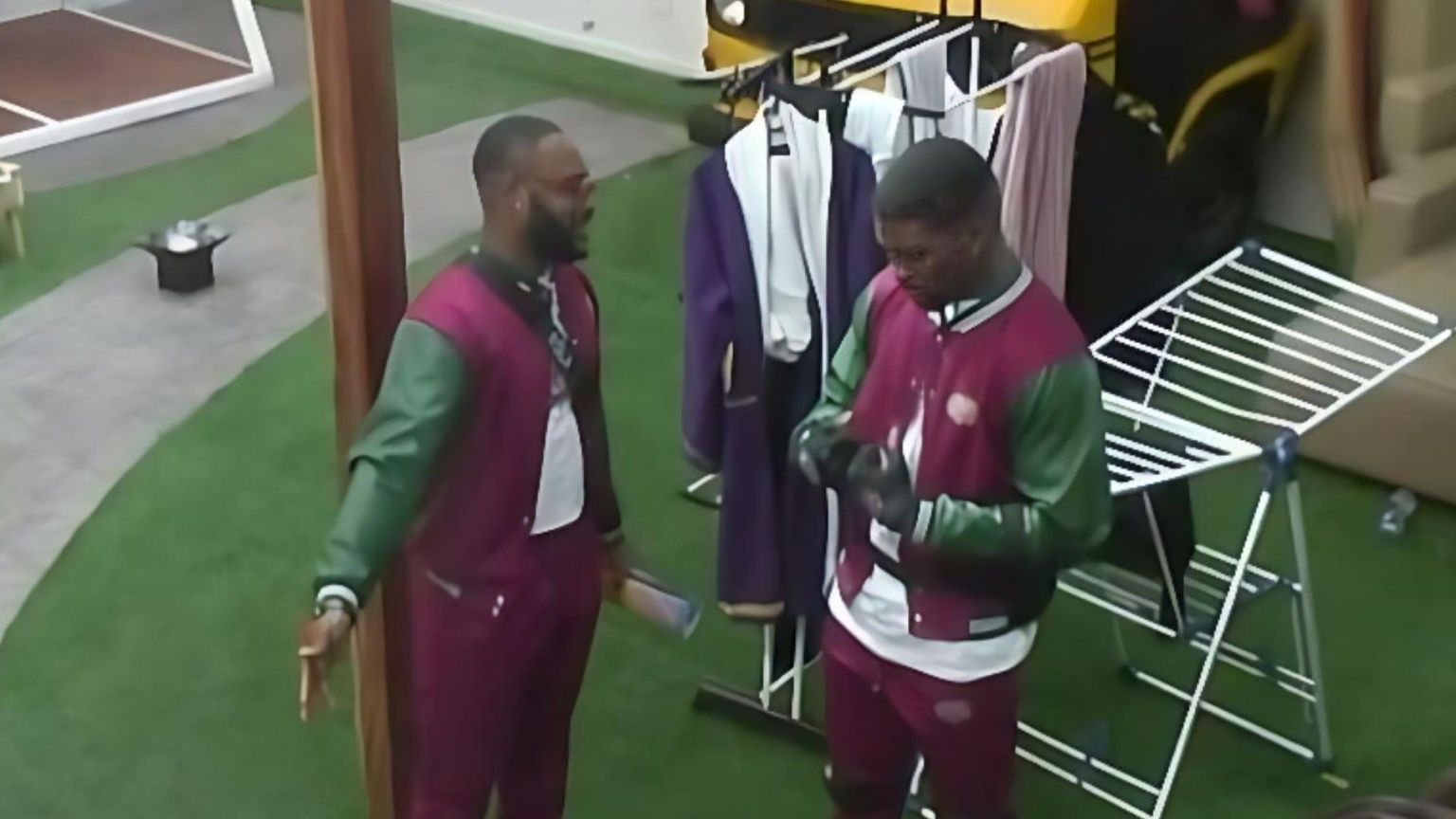 BBNaija Season 10 housemates Kola and Bright Morgan pictured outdoors during a task session, both dressed in matching maroon and green varsity-style outfits while engaging in conversation.