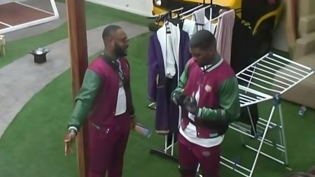 BBNaija Season 10 housemates Kola and Bright Morgan pictured outdoors during a task session, both dressed in matching maroon and green varsity-style outfits while engaging in conversation.