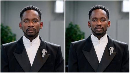 Mr Eazi poses in a black tuxedo jacket with a white high-neck shirt and a silver brooch, standing indoors against a blurred background.
