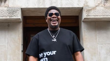 Nigerian comedian and activist Mr. Macaroni smiles brightly in a black T-shirt with bold white text, layered silver necklaces, and dark sunglasses, standing in front of a stone building entrance.