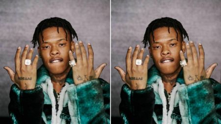 South African rapper Nasty C in a green fur coat, showing his rings and tattoos with both hands raised, looking straight at the camera with a slight lip bite.