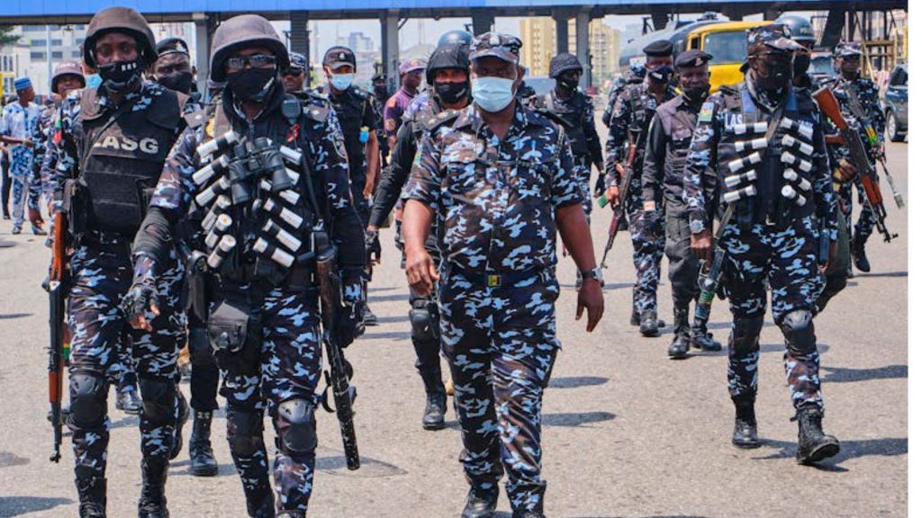 group of Nigerian police officers in full camouflage gear, helmets, and bulletproof vests march together on a city street, some carrying rifles and tear gas canisters strapped to their vests.