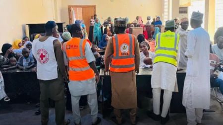 Nigerian evacuees from Sudan sit inside a hall as officials in Red Cross, NEMA, and SEMA reflective vests attend to them during their return and documentation process in Kano.