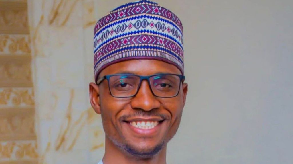 A smiling man wearing glasses and a patterned traditional Hausa/Fulani cap, dressed in a light outfit, standing against a neutral background.