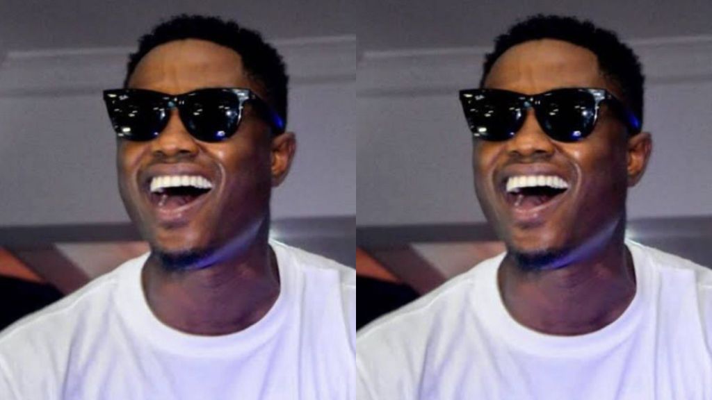 Nigerian rapper Vector captured in a mirrored photo effect, smiling widely in dark sunglasses and a white T-shirt during an event.