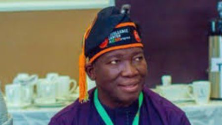 Nigerian man Wasiu Afonabi wears a traditional cap with orange and green details while seated indoors with cups and saucers in the background.