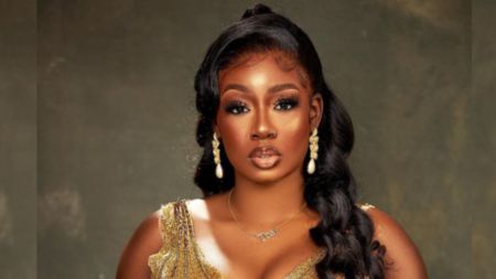 BBNaija star Tolani Baj poses in a gold embellished dress, wearing drop pearl earrings and a necklace with her name, with her long wavy hair styled to the side.