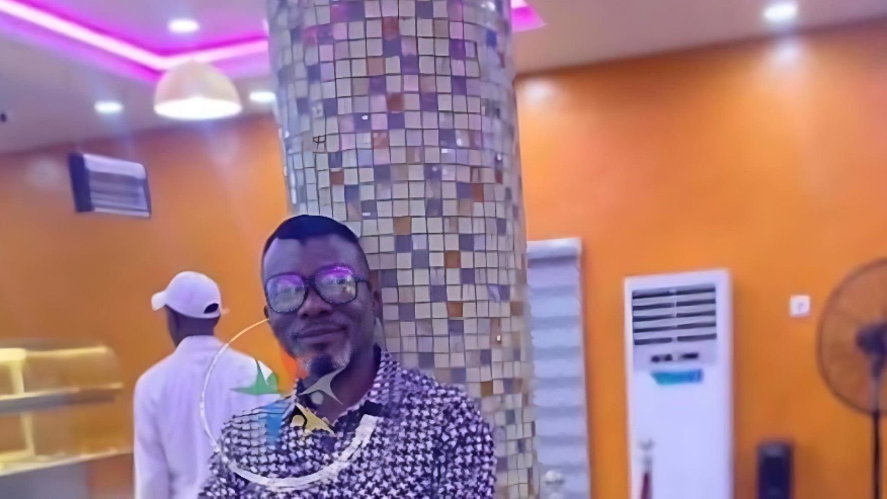 Nigerian man Alex, who was tragically killed by gunmen at a nightclub in Lagos, poses indoors wearing glasses and a patterned shirt, standing against a tiled pillar with an orange wall in the background.