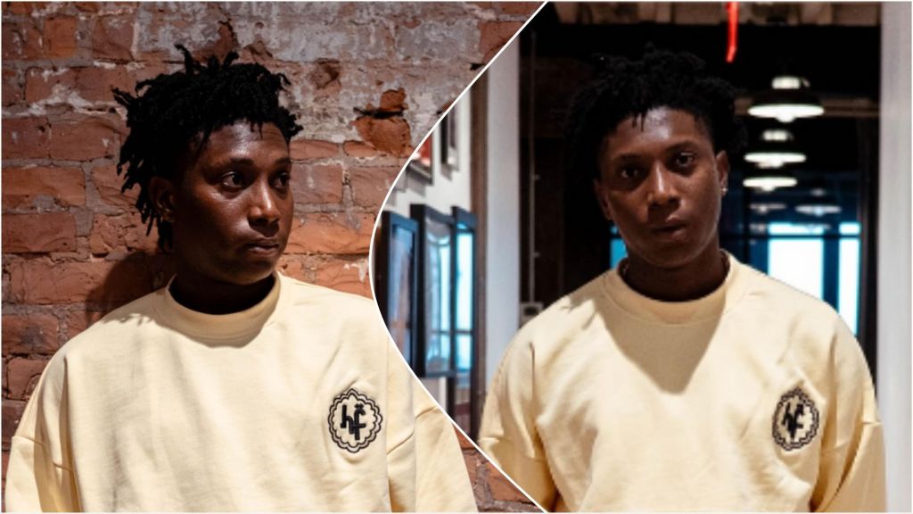 Nigerian singer Bella Shmurda poses indoors wearing a cream sweatshirt with a black emblem, against a brick wall and modern hallway background.
