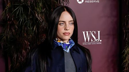 Singer Billie Eilish on the WSJ Magazine Innovator Awards 2025 red carpet, wearing a navy coat and patterned neck scarf, standing in front of a maroon WSJ backdrop.