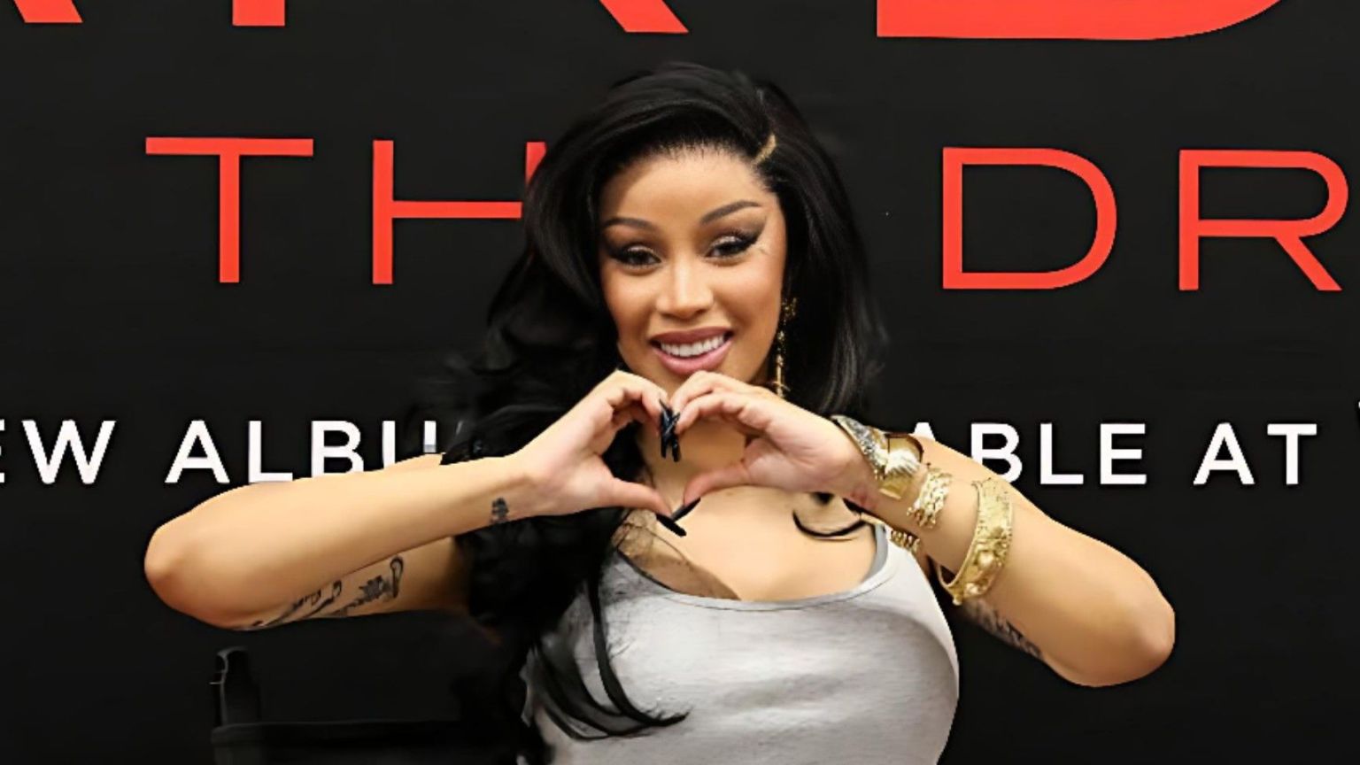 Cardi B smiles and forms a heart shape with her hands while posing in front of a black and red “Am I The Drama” album backdrop.