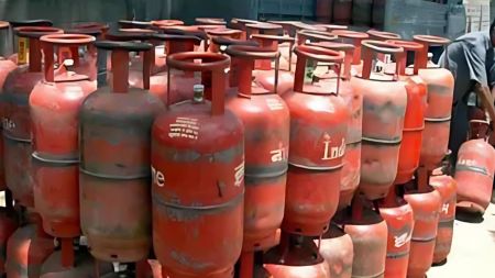 A row of red cooking gas cylinders arranged at a refilling station, with a worker seen handling one of the cylinders on the right side of the image.